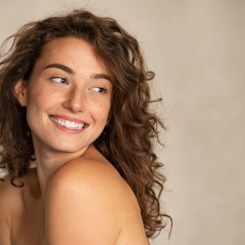 Portrait of smiling girl enjoying beauty treatment on beige background. Beautiful natural woman looking at copy space, spa and wellness concept. Carefree laughing woman with bare shoulders isolated.