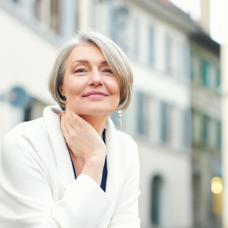 Outdoor portrait of beautiful and elegant middle age 55 - 60 year old woman, wearing white jacket, sitting on bench outside