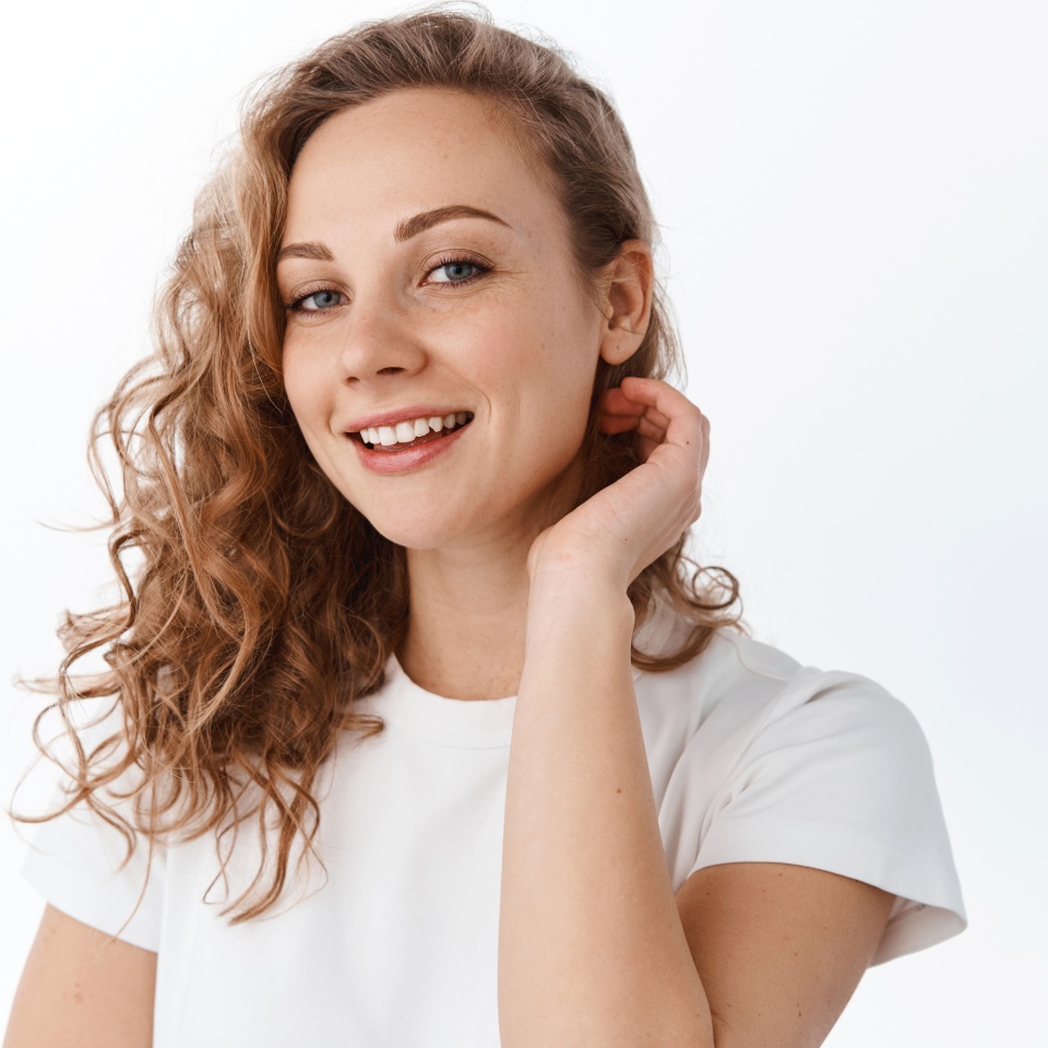 Young woman with curly blond hairdo, tuck hair behind ear and smile happy at camera, look natural and relaxed, standing over white background