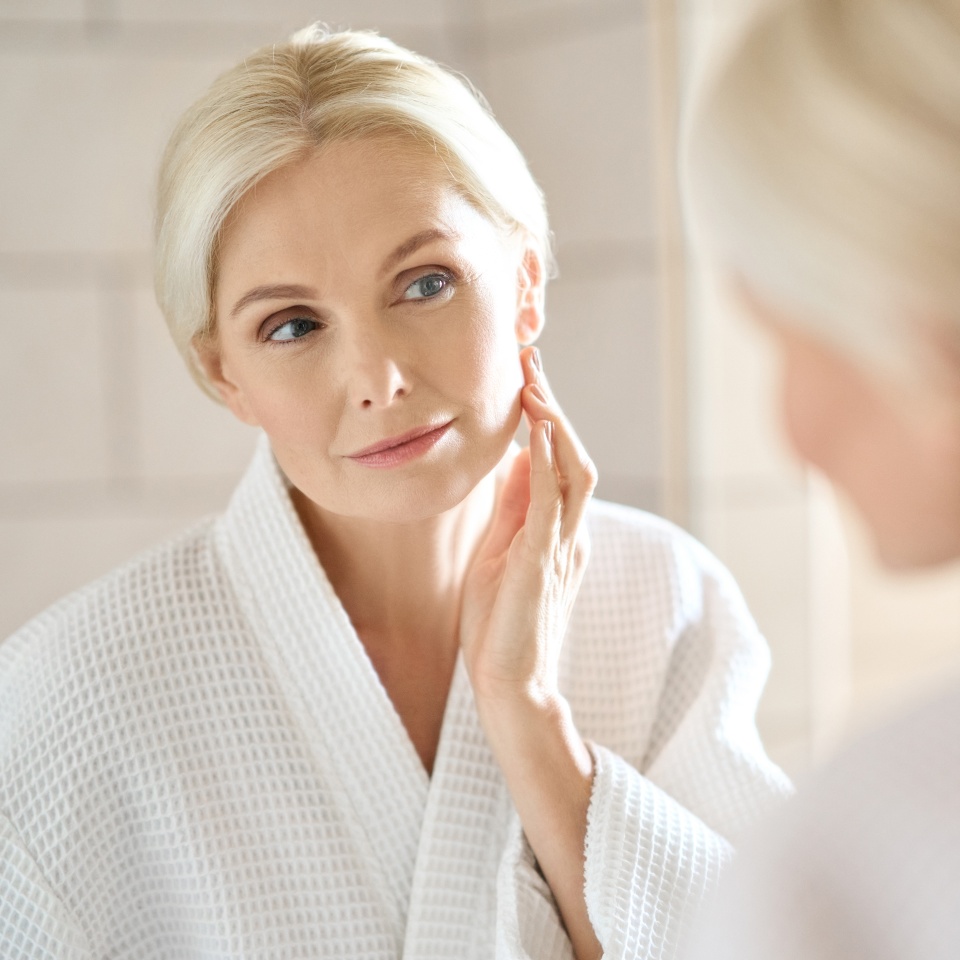 Headshot of gorgeous mid age adult 50 years old blonde woman standing in bathroom after shower touching face, looking at reflection in mirror doing morning beauty routine. Older skin care concept.