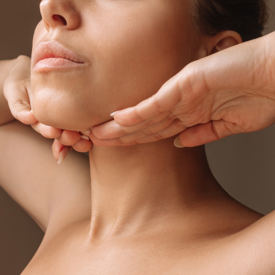 Cropped shot of young caucasian woman touching under the chin with hands massaging her face on dark brown background. Rejuvenation, facelift, facefitness. Exercises from the second chin, pelican neck