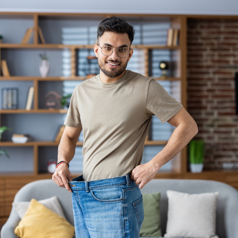 Portrait of a young smiling Indian man standing at home in front of the camera wearing jeans and showing the result of diet, exercise and weight loss.