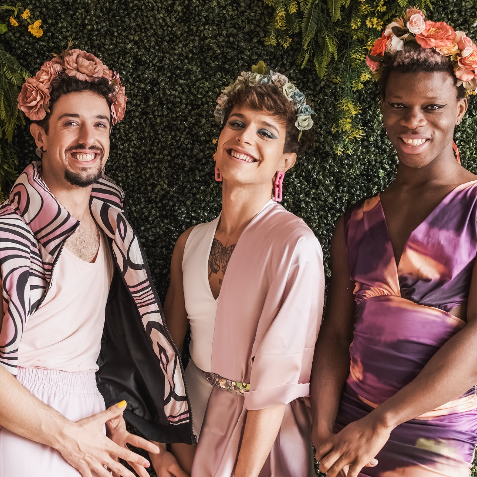 Three people wearing floral headpieces and colorful clothing stand smiling in front of a green leafy background.