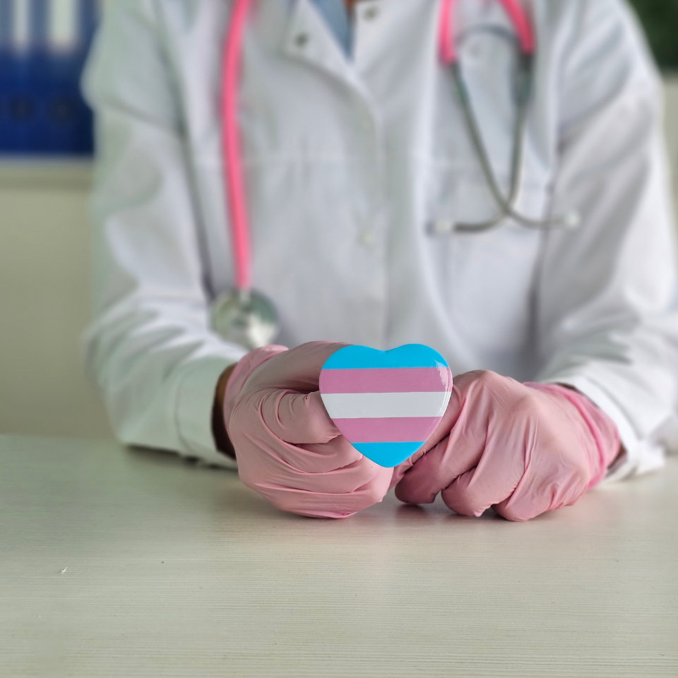 A person in a lab coat and pink gloves holds a heart-shaped object with the transgender pride flag colors; a stethoscope is draped around their neck.