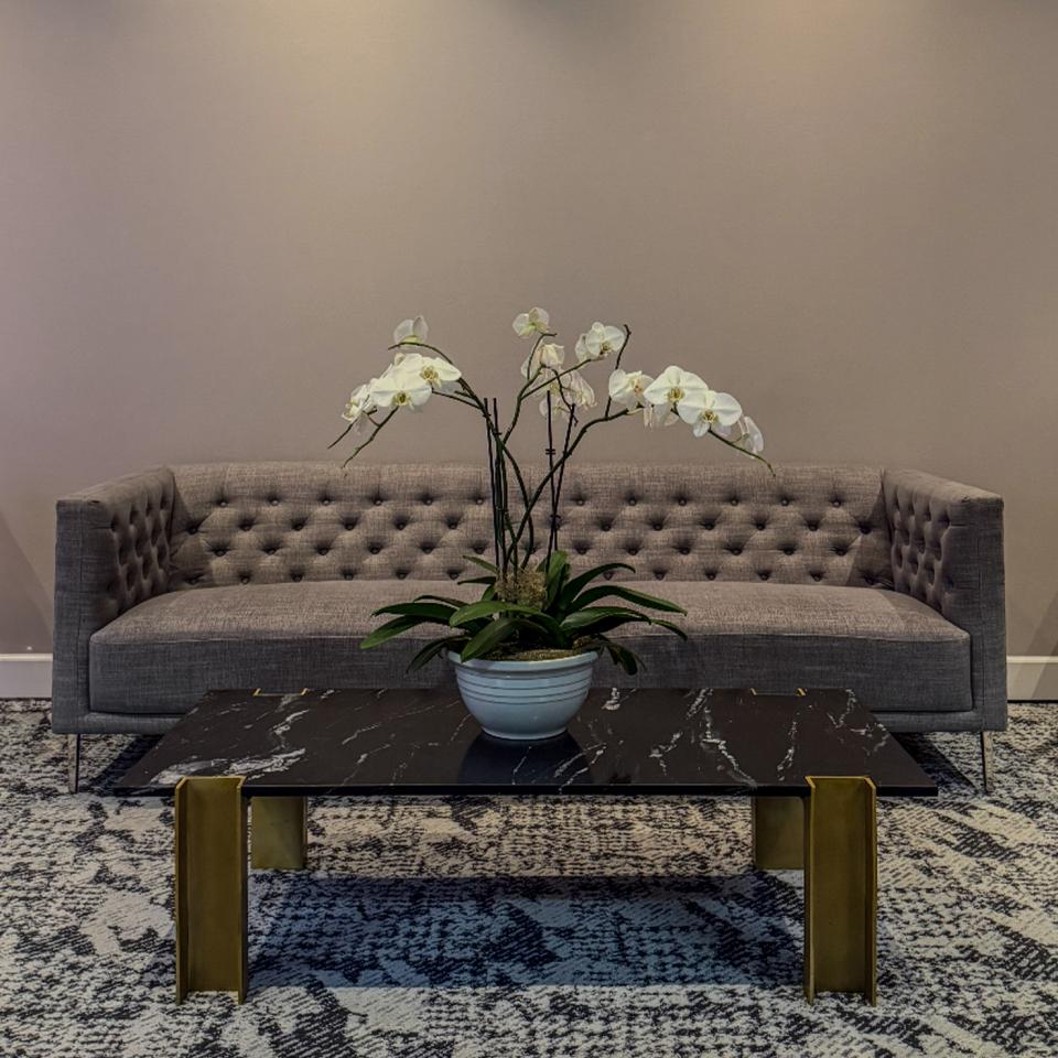 A modern lounge area with a gray sofa, two white armchairs, a marble coffee table with a plant and decorative items, and patterned carpeting.
